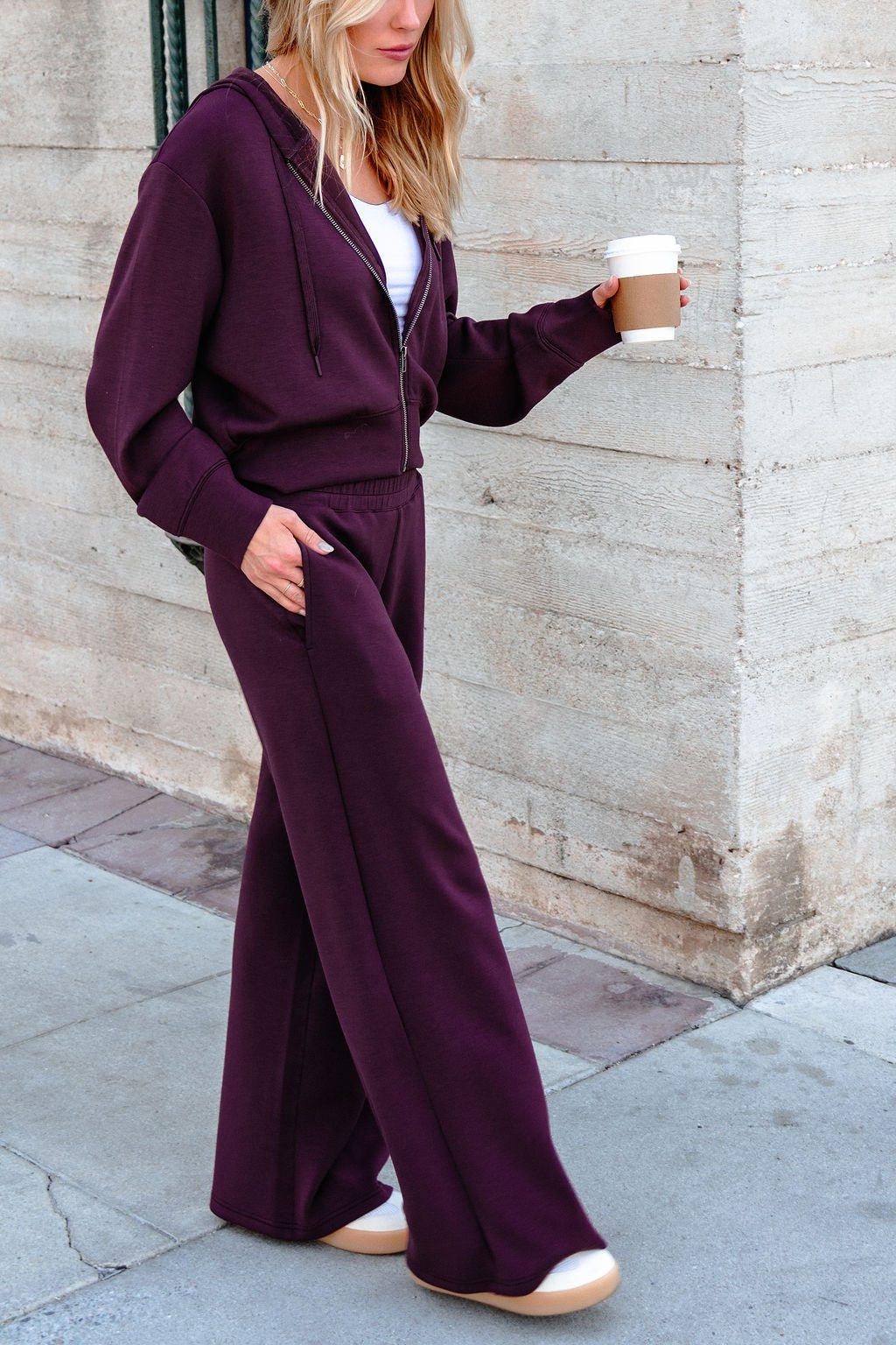 A woman in Vintage Havana Merlot Cloud Fleece Flare Pants, holding coffee, walks past a light stone wall.