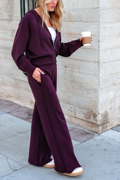 A woman in Vintage Havana Merlot Cloud Fleece Flare Pants, holding coffee, walks past a light stone wall.