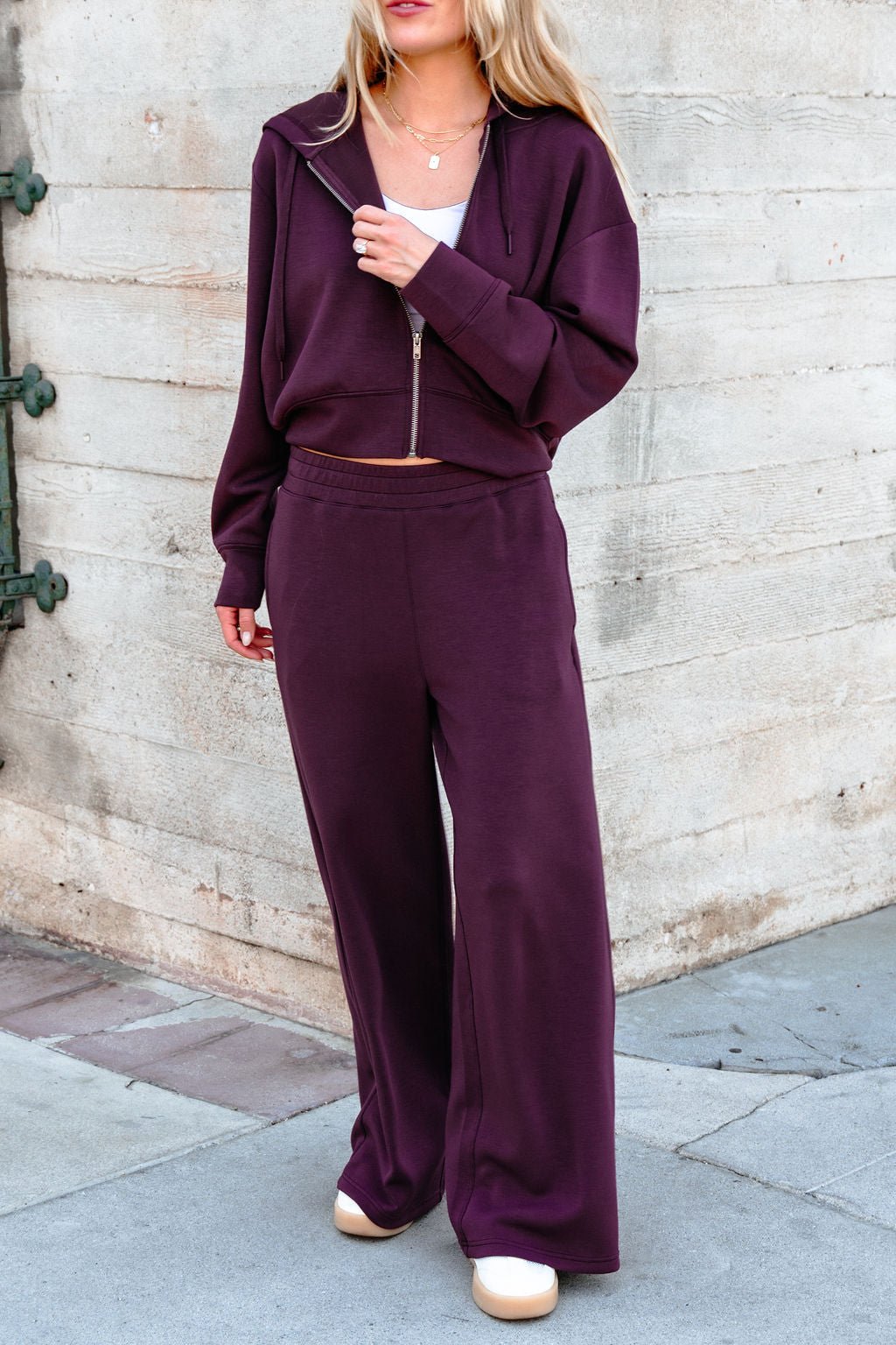 Woman wearing a maroon zip-up hoodie and Vintage Havana Merlot Cloud Fleece Flare Pants, standing by a concrete wall.