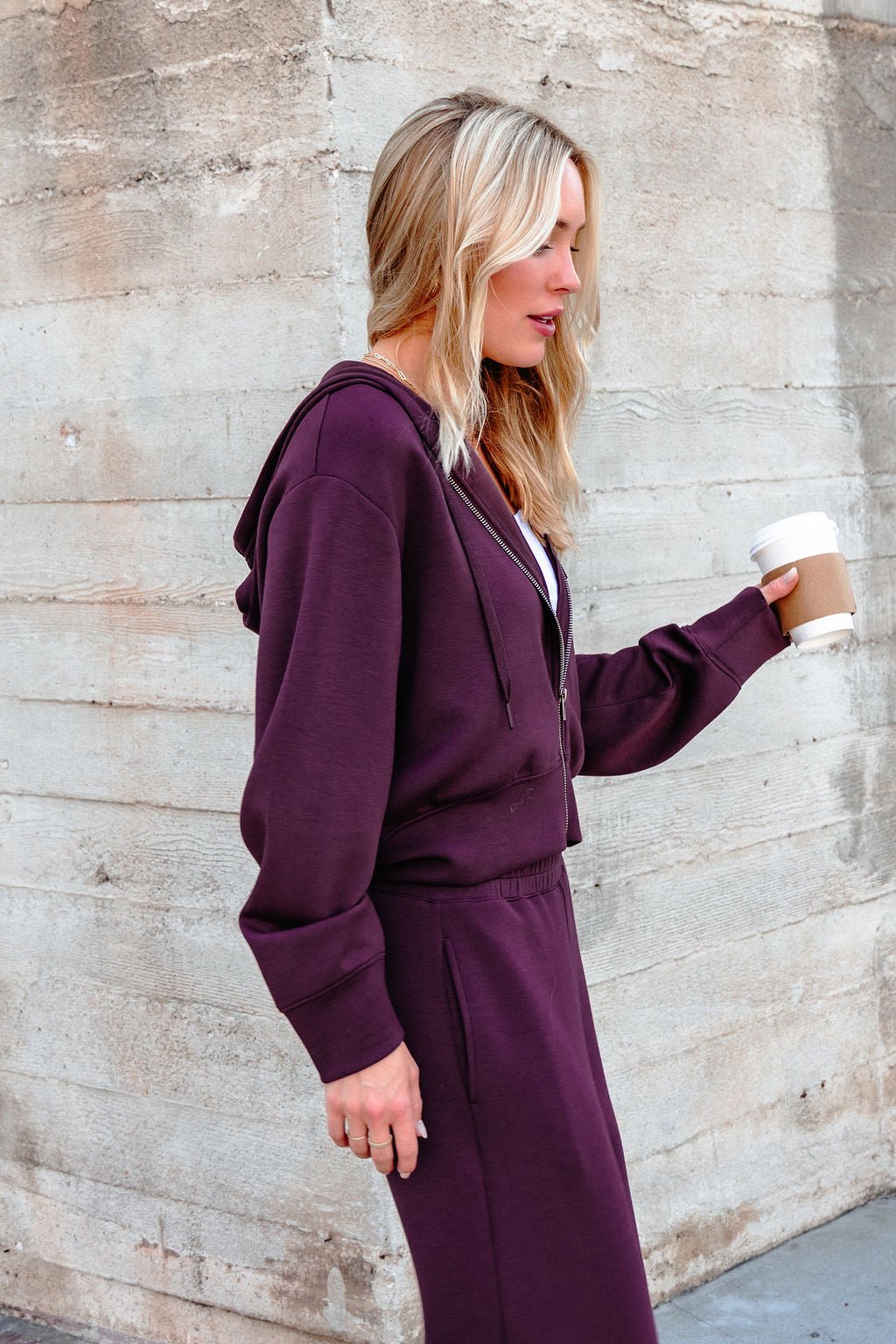 A woman in a Vintage Havana Merlot Cloud Fleece Zip Up Hoodie holds a coffee cup while walking past a concrete wall.