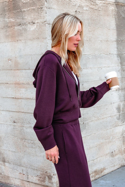 A woman in a Vintage Havana Merlot Cloud Fleece Zip Up Hoodie holds a coffee cup while walking past a concrete wall.