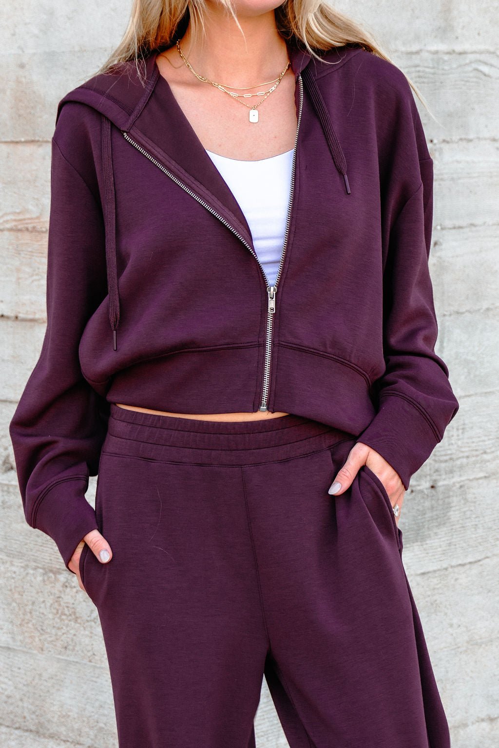 Woman in a Vintage Havana Merlot Cloud Fleece Zip Up Hoodie with matching pants, white top, and gold necklaces by a concrete wall.