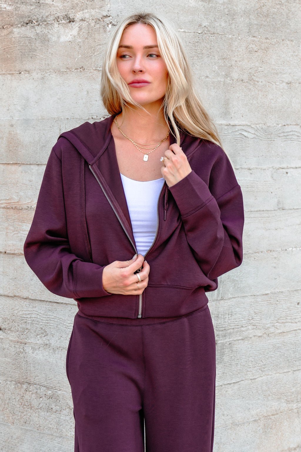Woman with long blonde hair wearing a Vintage Havana Merlot Cloud Fleece Zip Up Hoodie, standing by a textured wall.