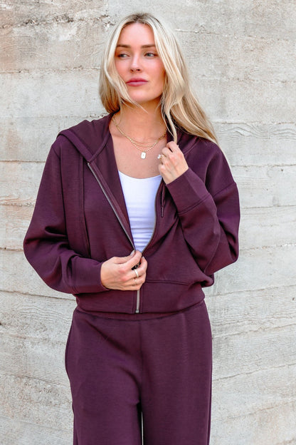 Woman with long blonde hair wearing a Vintage Havana Merlot Cloud Fleece Zip Up Hoodie, standing by a textured wall.