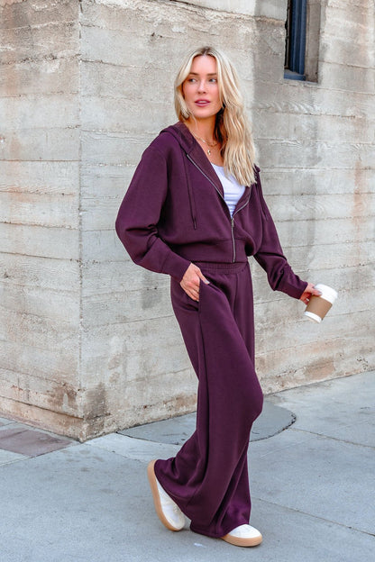 A woman in a Vintage Havana Merlot Cloud Fleece Zip Up Hoodie holds a coffee cup while walking by a concrete wall.