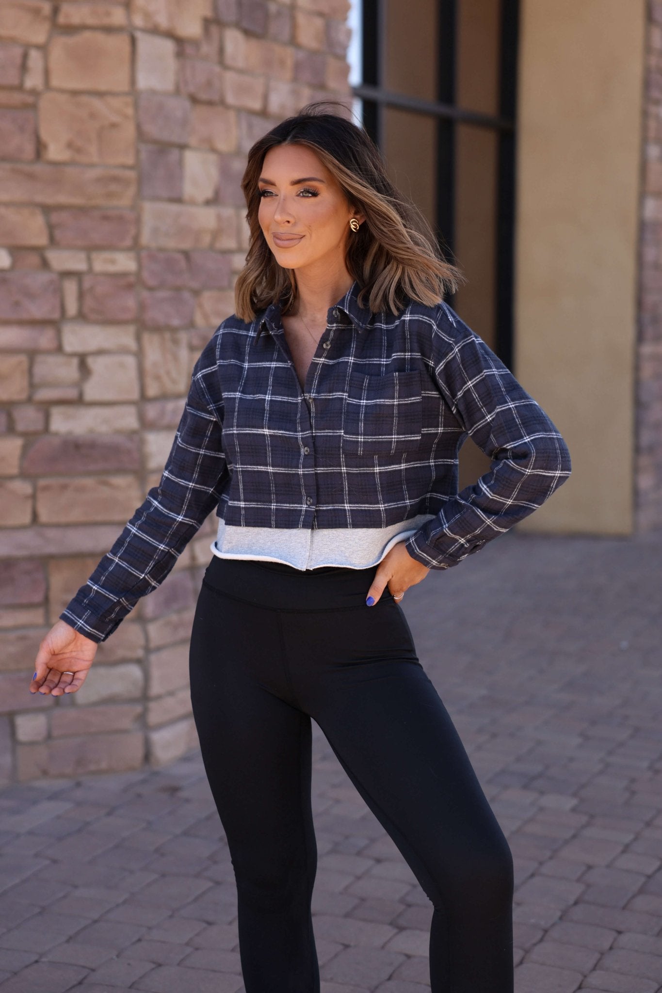A woman smiles outdoors in a Vintage Havana Midnight Plaid Flannel Shirt and black leggings, hand on hip.