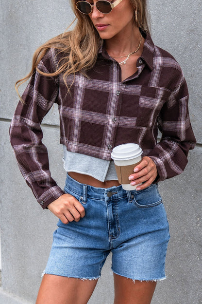 Woman in sunglasses and denim shorts wears the Vintage Havana Mocha Plaid Flannel Shirt, standing by a gray wall with a coffee cup.