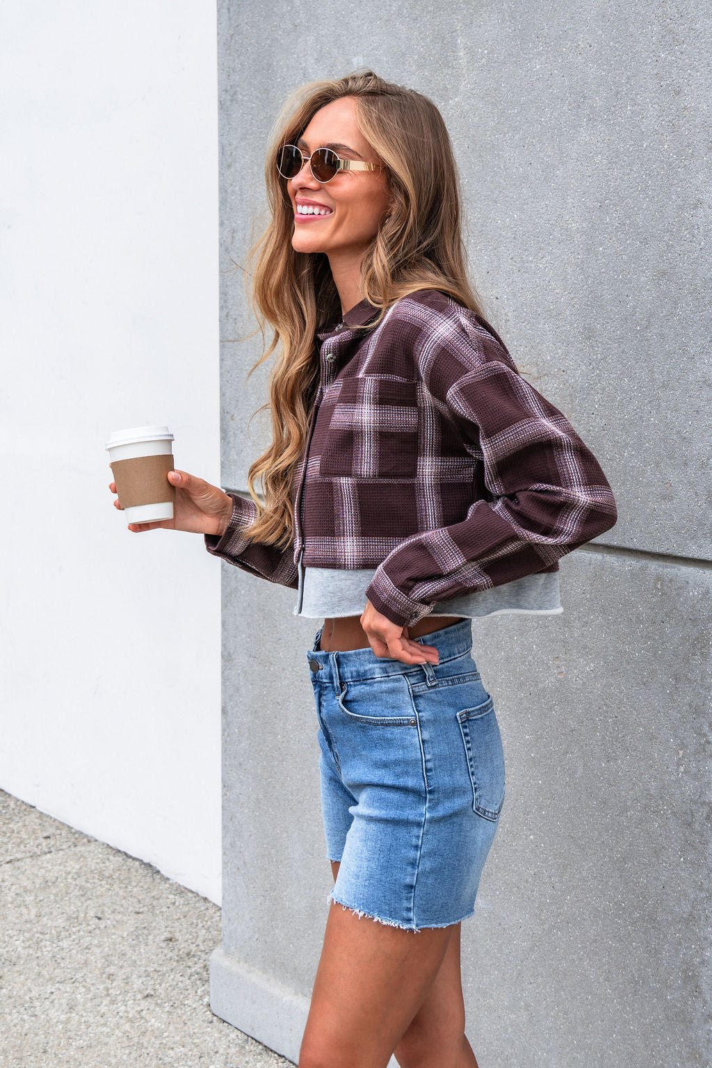 Woman in sunglasses wears the Vintage Havana Mocha Plaid Flannel Shirt, holding coffee and smiling by a gray wall.