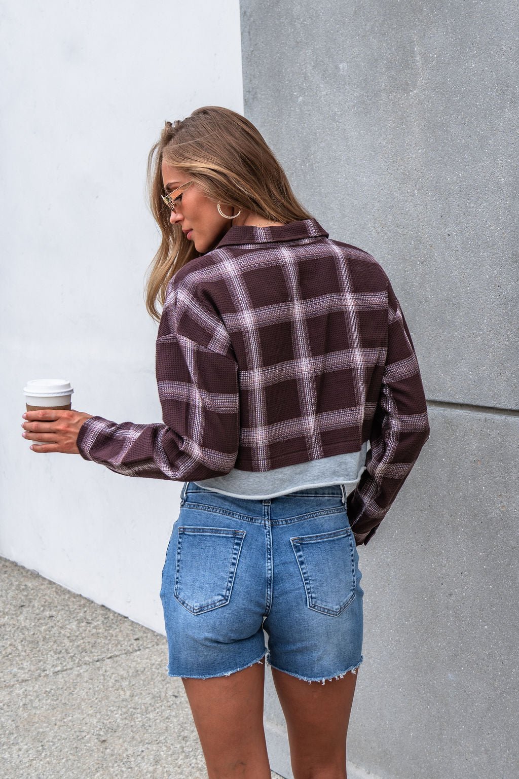 Woman with long hair wears Vintage Havana Mocha Plaid Flannel Shirt, denim shorts, and holds coffee by a concrete wall, facing away.