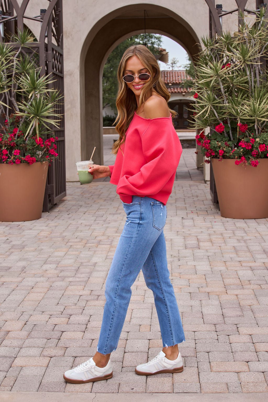 Woman in a Vintage Havana Off The Shoulder Pullover - Cherry, blue jeans, and white sneakers, smiling outdoors with a drink.