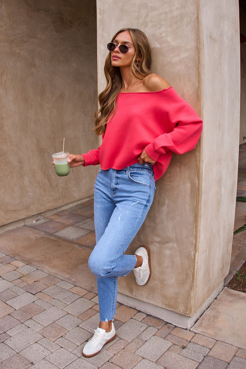 Woman in sunglasses wears the Vintage Havana Off The Shoulder Pullover in Cherry, blue jeans, and white sneakers, holding a drink.