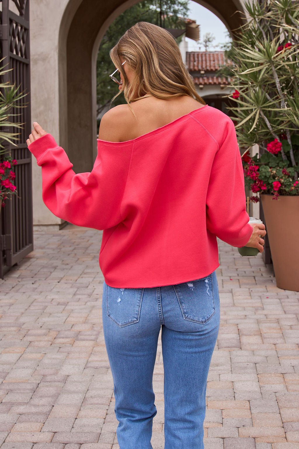 A woman in the Vintage Havana Off The Shoulder Pullover - Cherry and blue jeans stands outdoors, facing away with one arm raised.