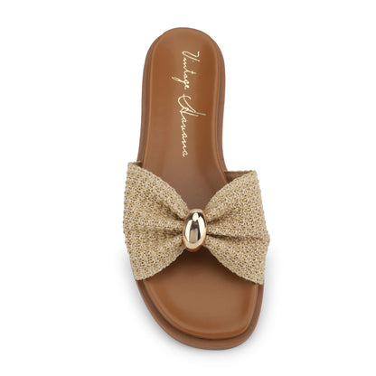 The Vintage Havana Yvette Beige Slide Sandals feature a tan woven strap with a bow, gold accent, flat sole, and open toe.