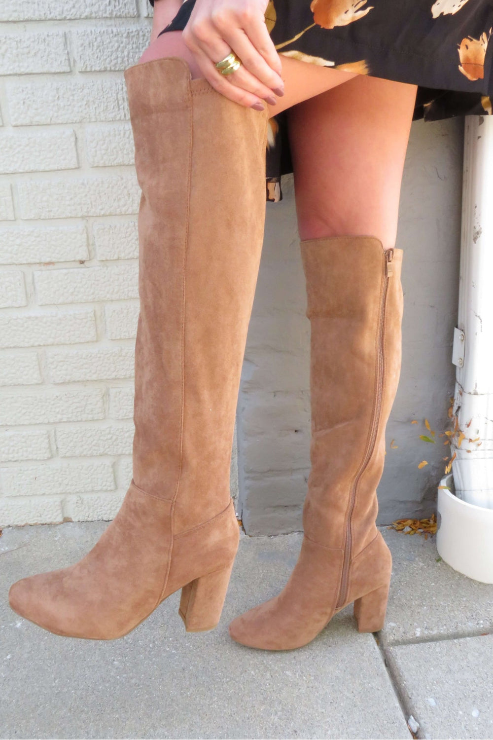 A person stands by a brick wall wearing Whisky Camel Suede Heeled Knee High Boots - FINAL SALE and a black floral skirt.