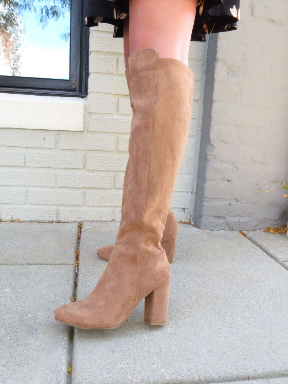 Someone wears Whisky Camel Suede Heeled Knee High Boots, standing on a sidewalk by a light brick wall.