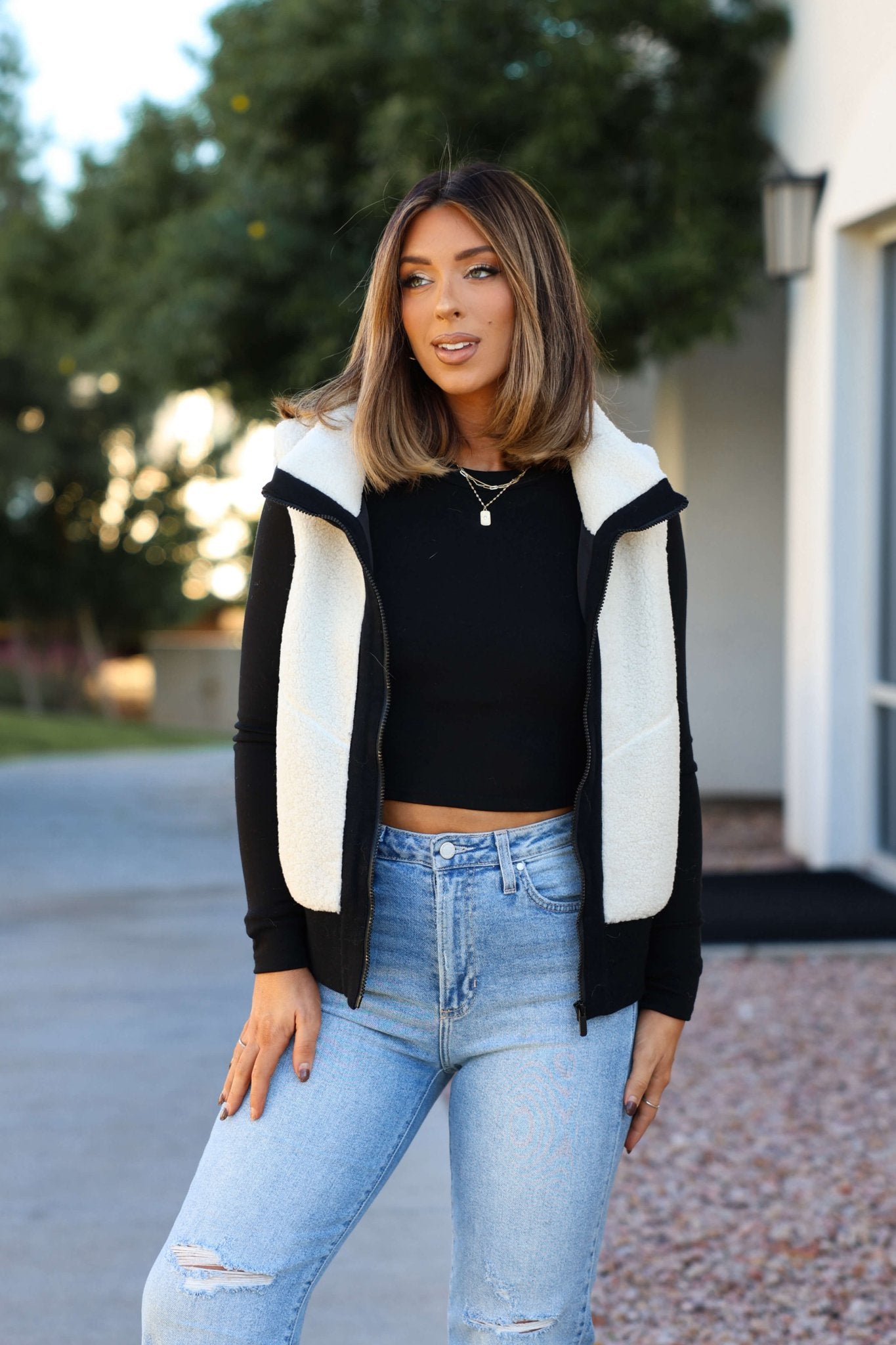 A woman with shoulder-length hair wears a black top, light blue jeans, and a white and black color block sherpa zip vest outdoors.