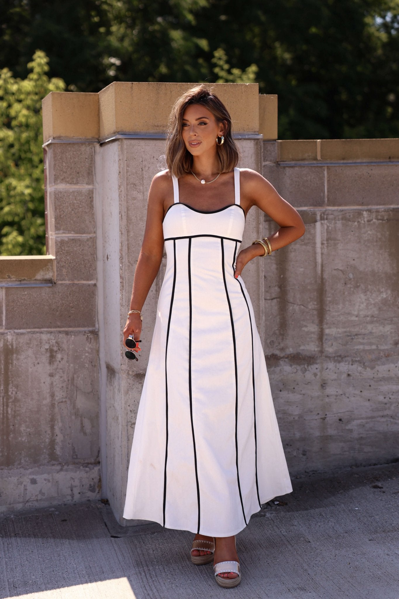 Woman in a White and Black Trim Detail Maxi Dress stands outdoors by a stone wall, holding sunglasses and wearing heels.
