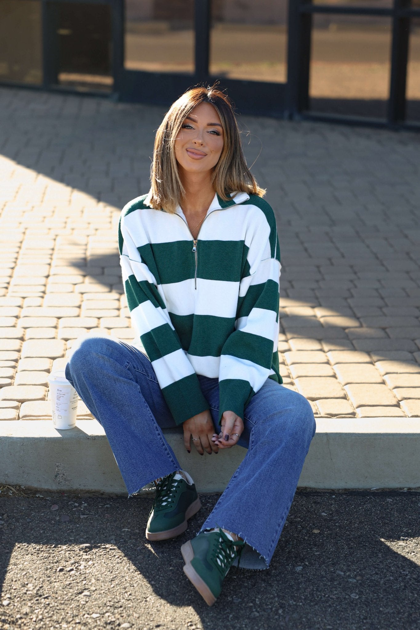 A woman in a White and Green Varsity Stripe Half Zip Rugby Pullover and blue jeans sits on a curb, smiling with a drink can beside her.