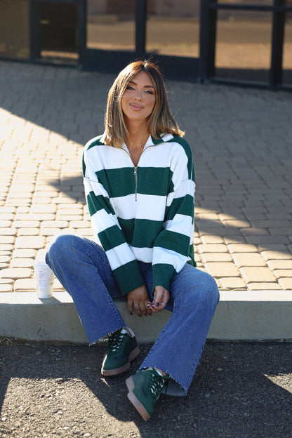 A woman in a White and Green Varsity Stripe Half Zip Rugby Pullover and blue jeans sits on a curb, smiling with a drink can beside her.