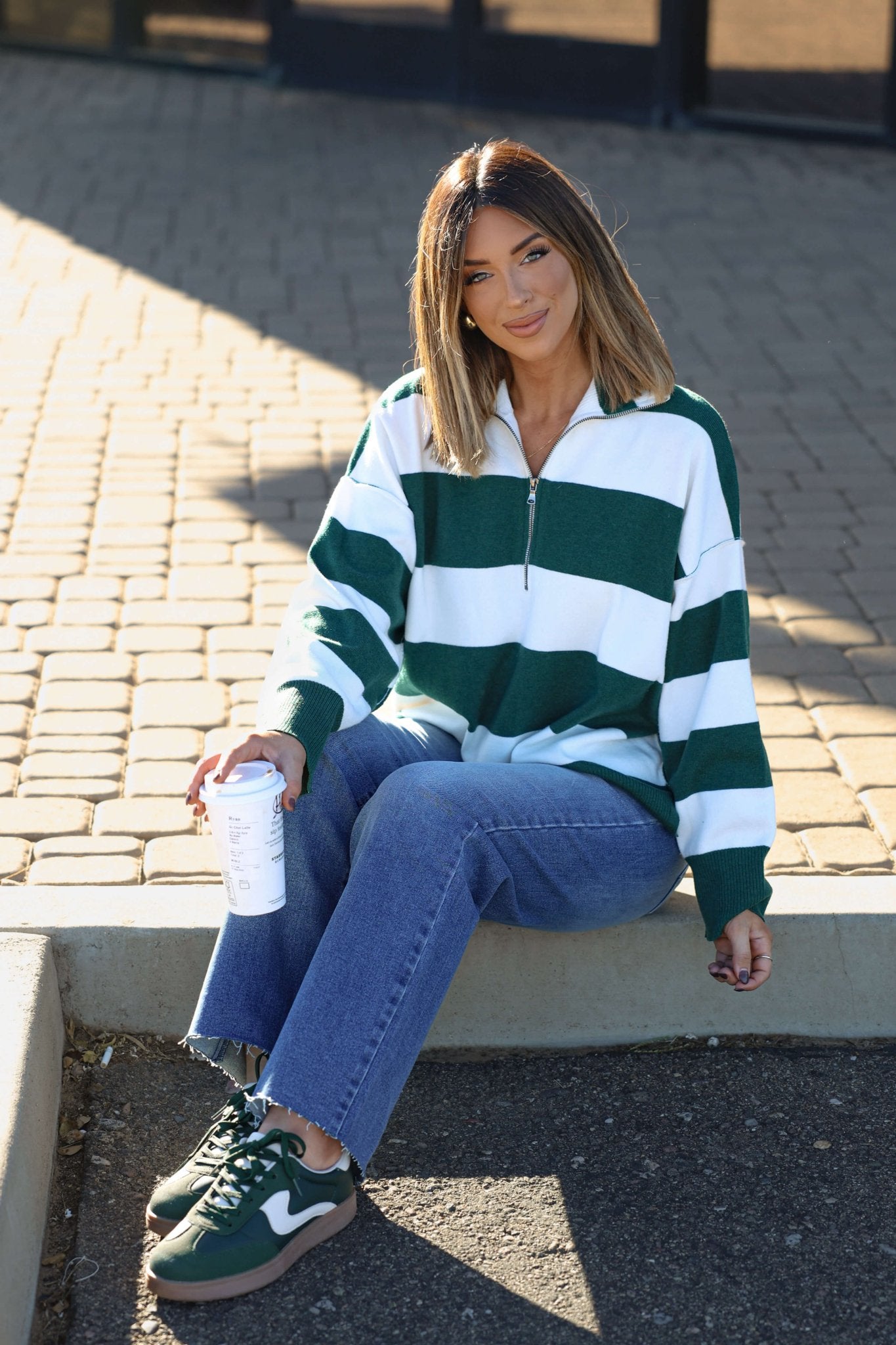 Woman outdoors in a white and green varsity stripe half zip rugby pullover, blue jeans, green sneakers, holding a coffee cup.