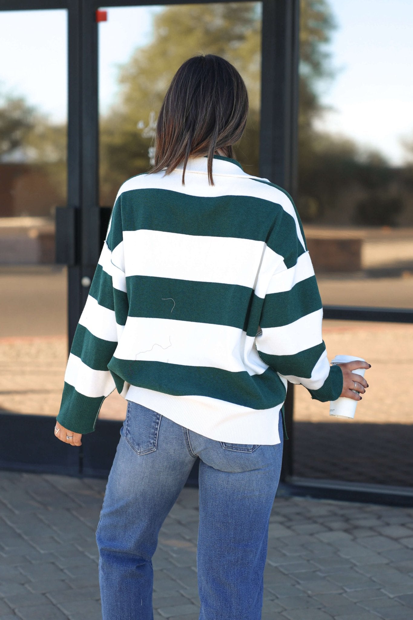 A woman in a White and Green Varsity Stripe Half Zip Rugby Pullover and jeans stands outside holding a cup, facing away.