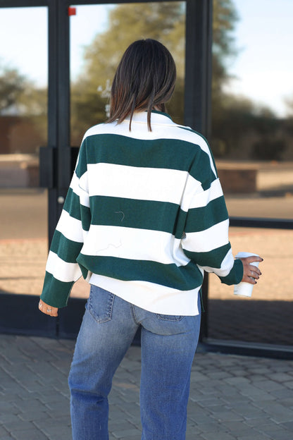 A woman in a White and Green Varsity Stripe Half Zip Rugby Pullover and jeans stands outside holding a cup, facing away.