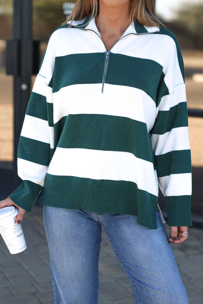 A woman in a White and Green Varsity Stripe Half Zip Rugby Pullover and blue jeans holds a coffee cup outdoors.