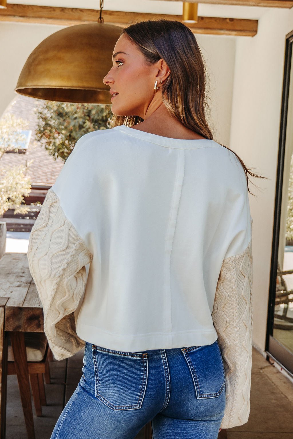 A woman with long hair wears the White Cable Knit Sweater Sleeve Top indoors, facing away. FINAL SALE.