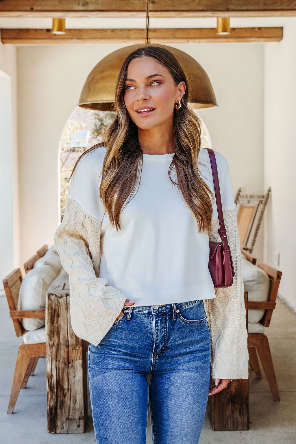 A woman smiles indoors in a White Cable Knit Sweater Sleeve Top, blue jeans, and a maroon bag over her shoulder.