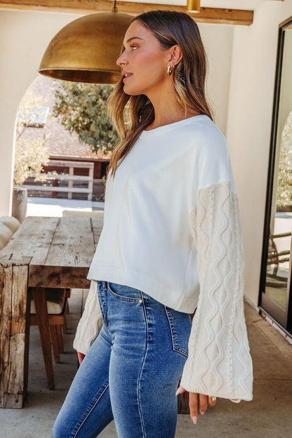 Woman in a White Cable Knit Sweater Sleeve Top and blue jeans stands indoors by a rustic wooden table.