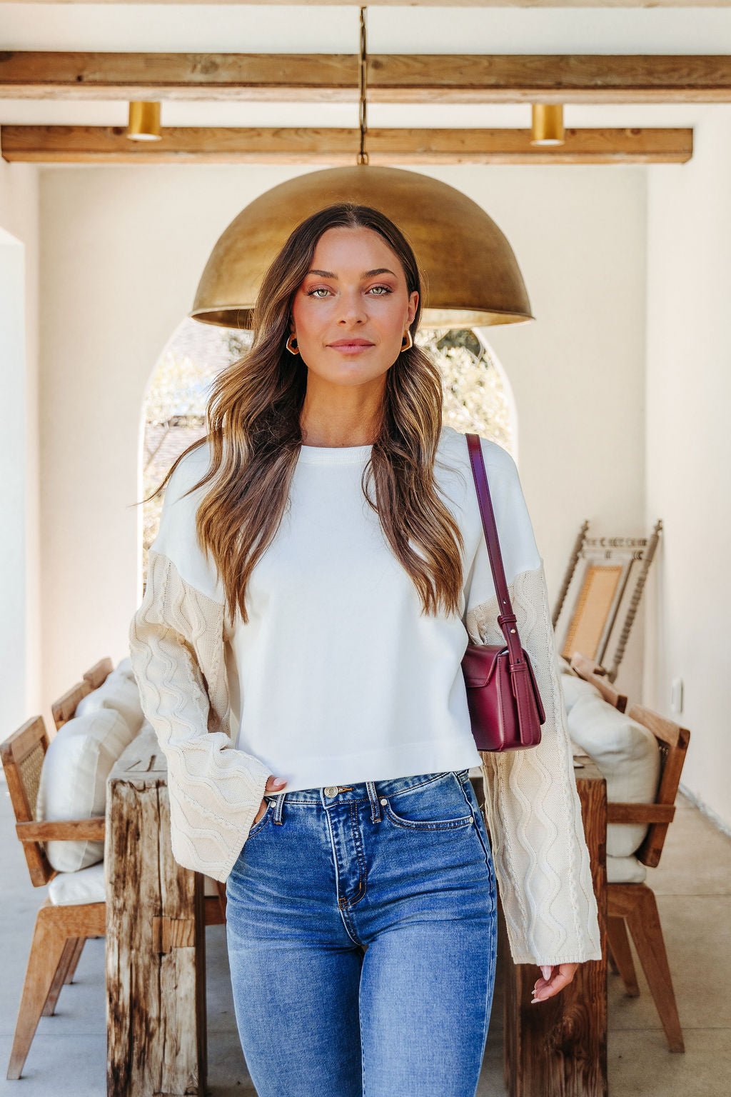 A woman with long wavy hair wears the White Cable Knit Sweater Sleeve Top in a stylish, rustic living space.