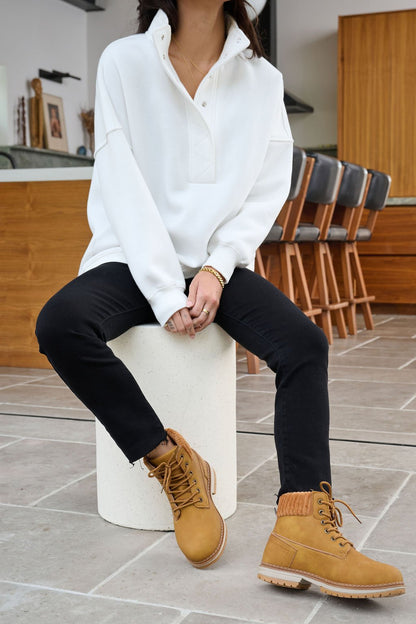 A woman in a White Collared Button Up Pullover Sweatshirt, black pants, and tan boots sits on a stool in a modern kitchen.