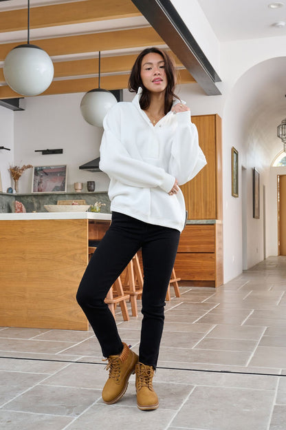 A woman in a Magnolia exclusive White Collared Button Up Pullover Sweatshirt, black pants, and tan boots poses in a modern kitchen.