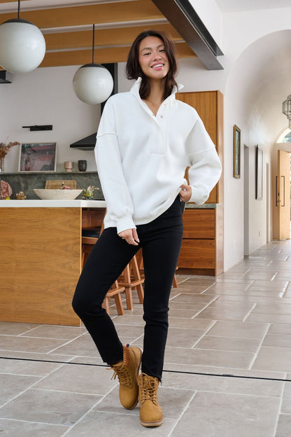 Woman smiling in a modern kitchen, wearing a Magnolia White Collared Button Up Pullover Sweatshirt, black pants, and tan boots.