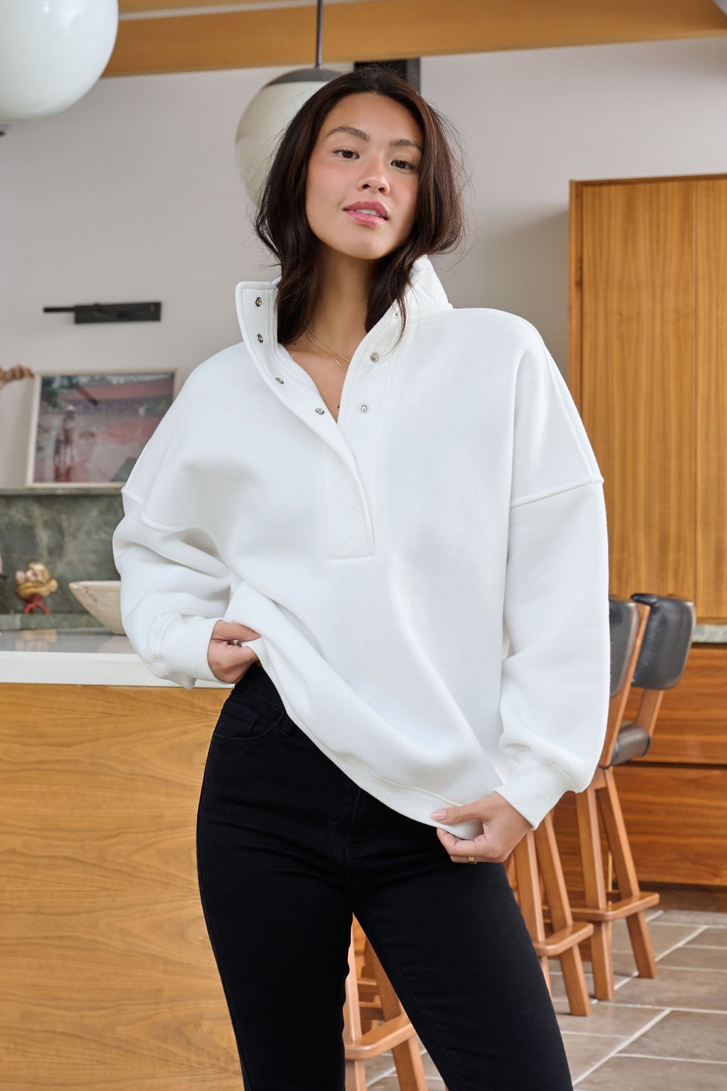 A woman indoors wears a White Collared Button Up Pullover Sweatshirt with black pants, holding the hem of her top.