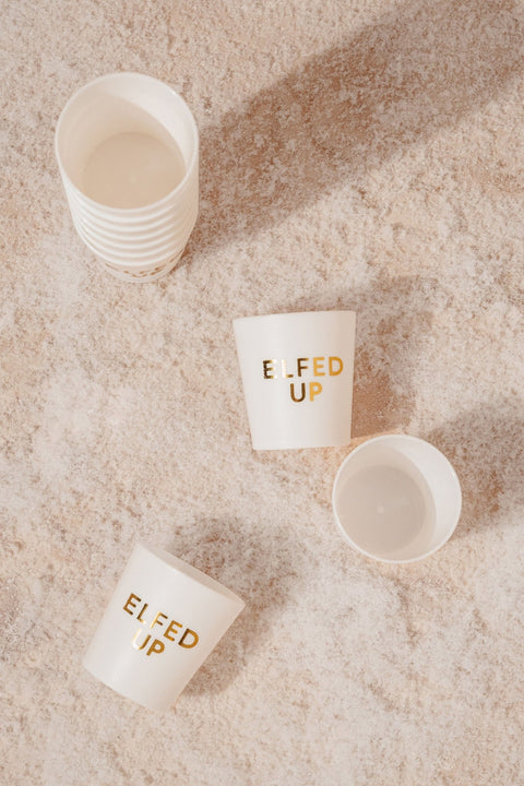 White Elfed Up Shot Cups - FINAL SALE shown on sand, two with “ELFED UP,” plus a festive stack and holiday shot cups nearby.