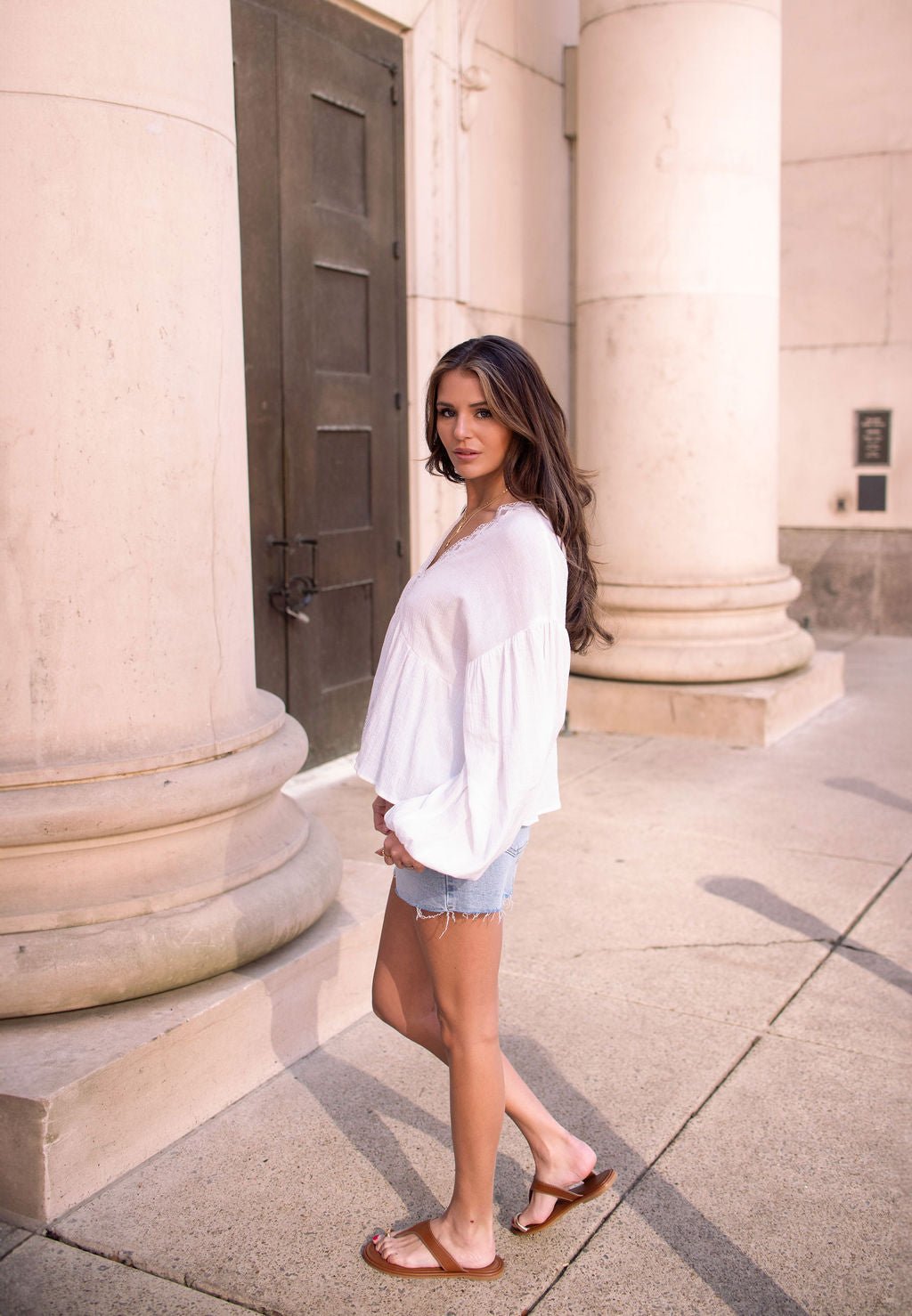 Woman wears the White Flowy Lace Trim Top - FINAL SALE with balloon sleeves and stands outdoors by columns, looking at the camera.