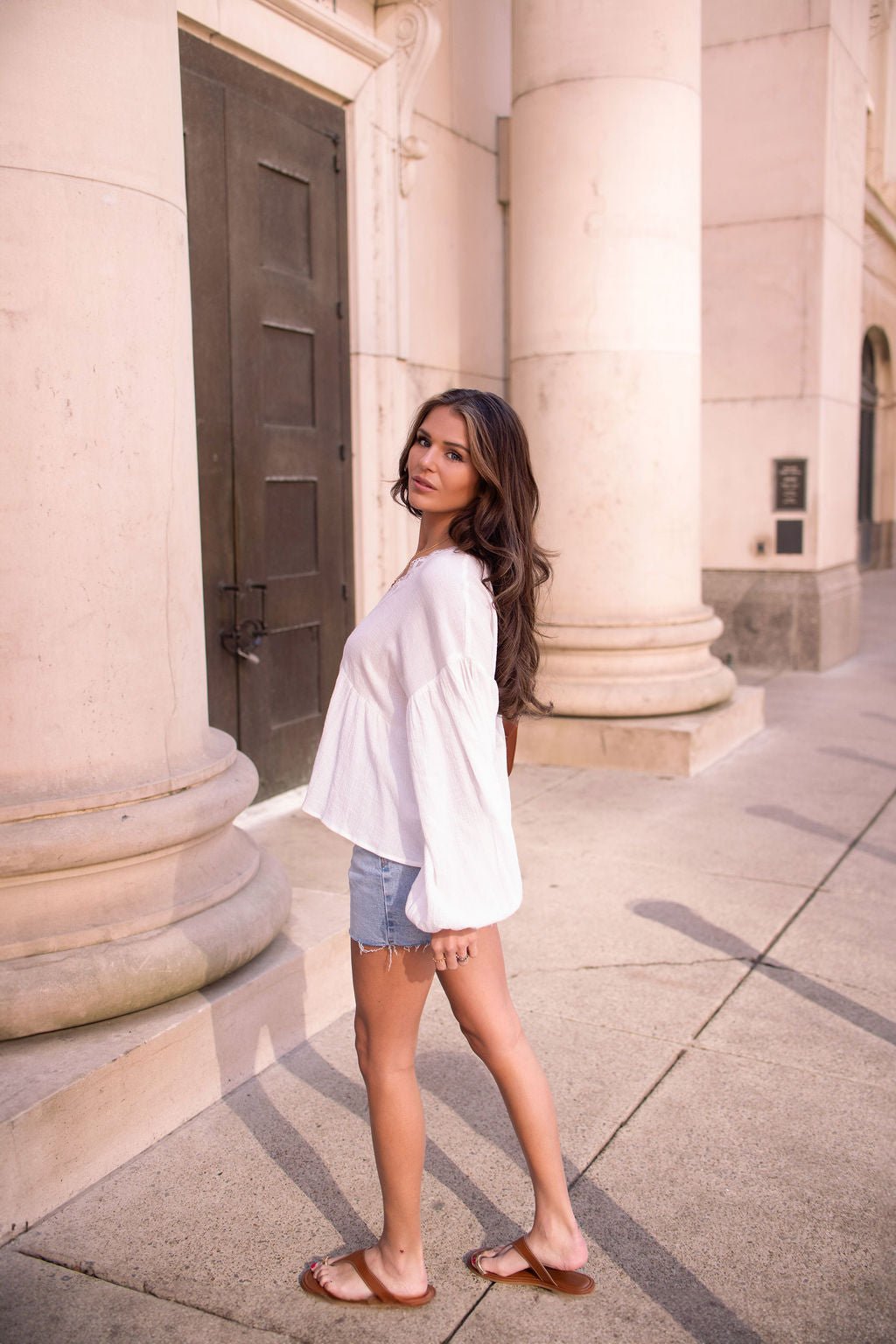 Wearing the White Flowy Lace Trim Top - FINAL SALE, denim shorts, and sandals, she stands by columns, looking over her shoulder.