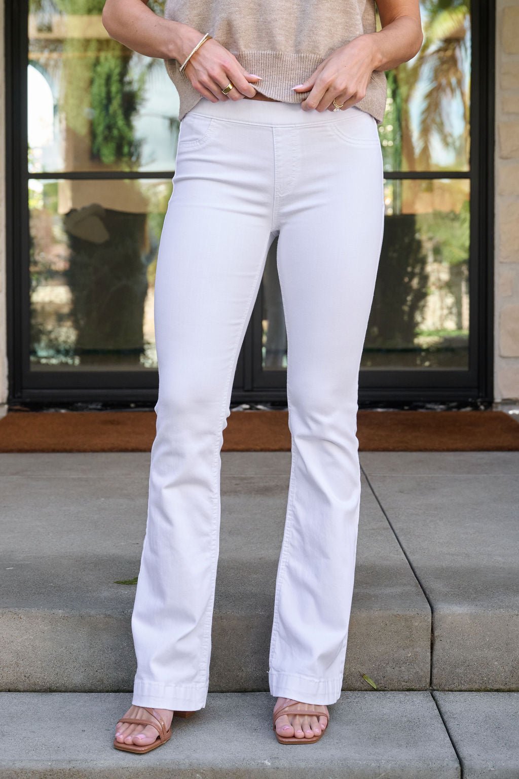 Woman in White Mid Rise Flare Pull-On Jeggings and sandals stands on outdoor steps by a glass door, upper body not shown.