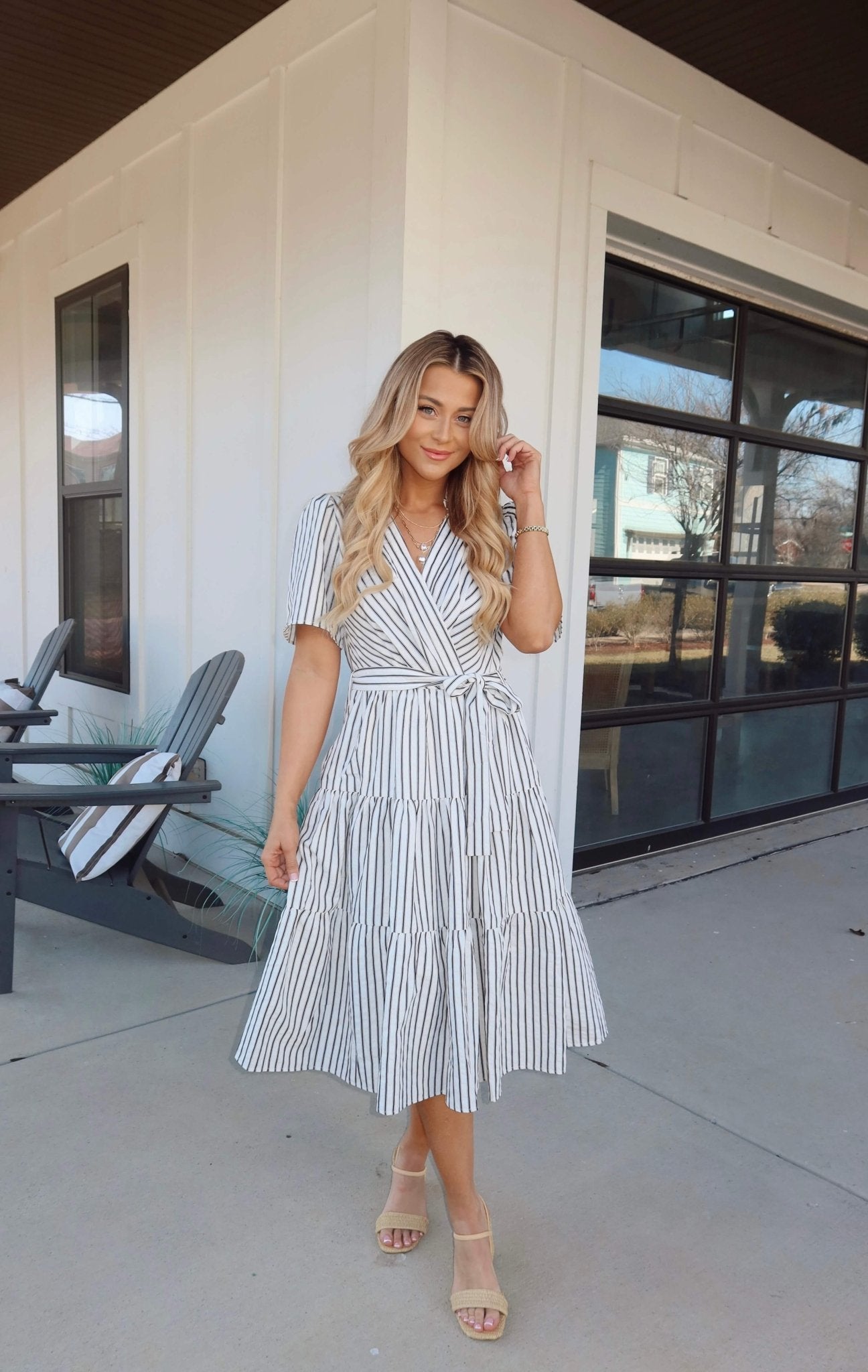 Woman in a White Stripe Surplice Tiered Midi Dress stands outside by a modern building with black-framed windows and gray chairs.