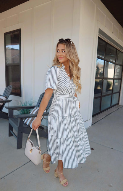 Woman in a White Stripe Surplice Tiered Midi Dress, beige handbag, sandals, and sunglasses stands outside a modern building.