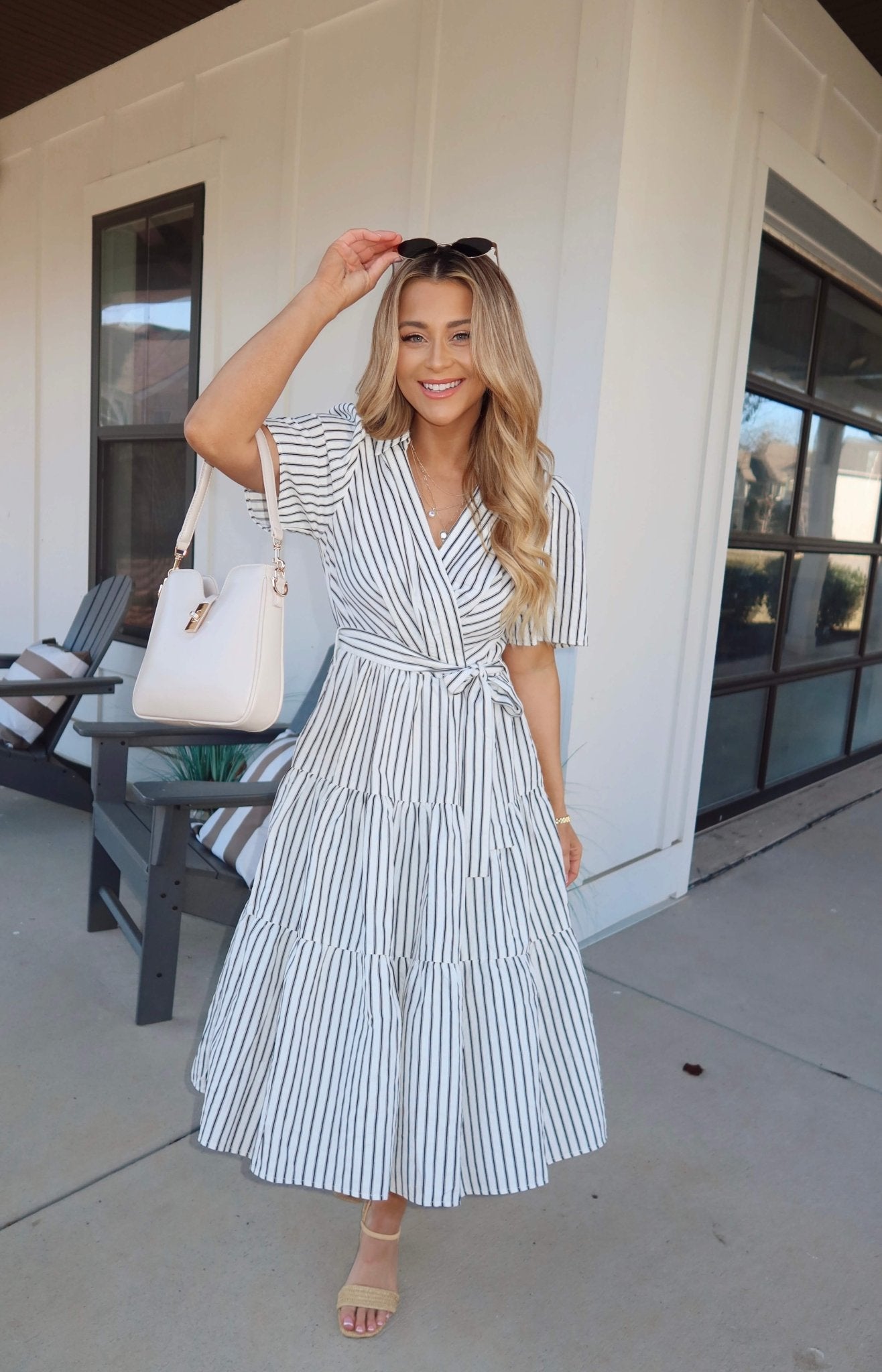 Woman in a White Stripe Surplice Tiered Midi Dress holds sunglasses and a white bag, smiling outside a modern building.