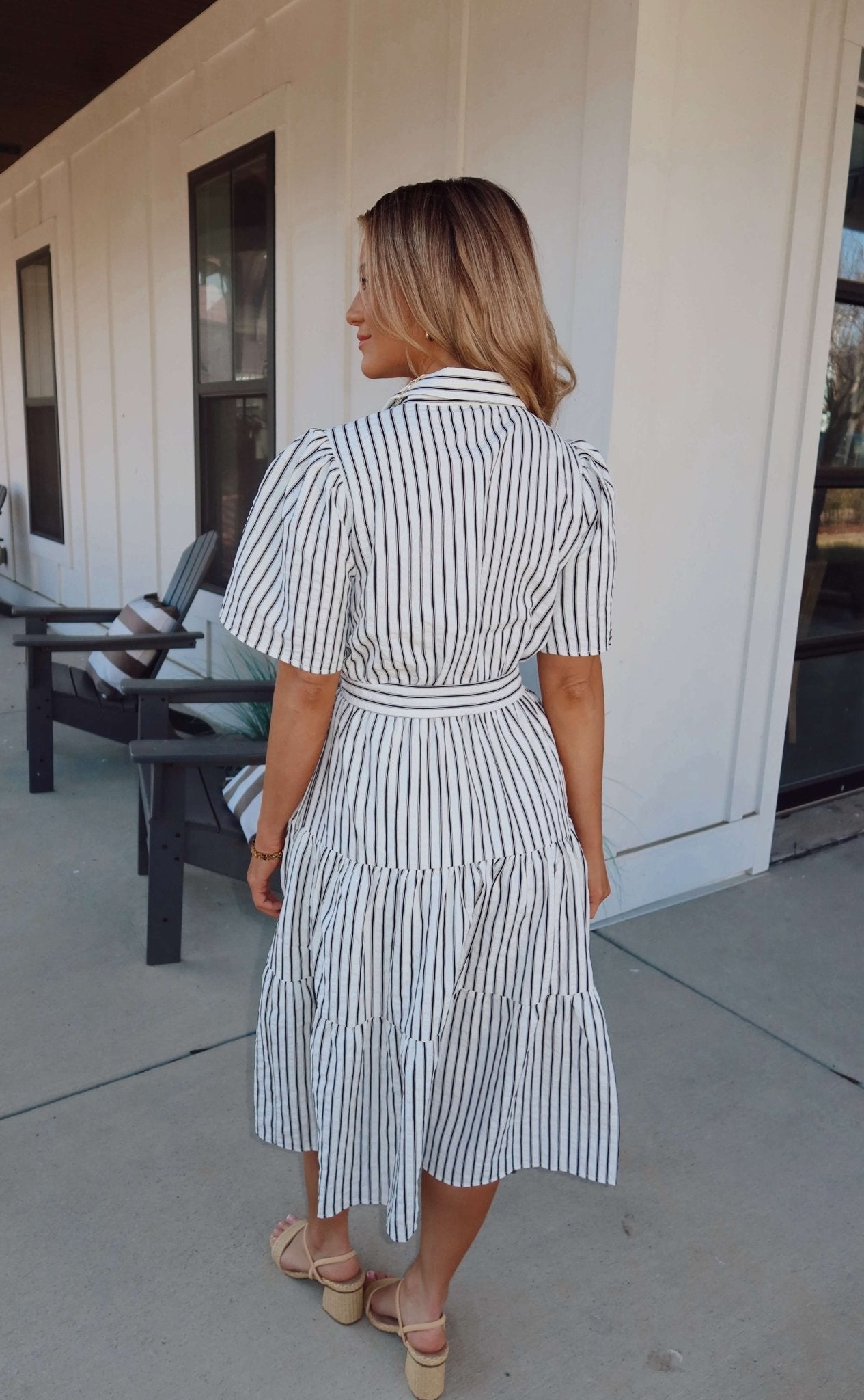 Woman in a White Stripe Surplice Tiered Midi Dress with black stripes stands outside near chairs, facing away from the camera.