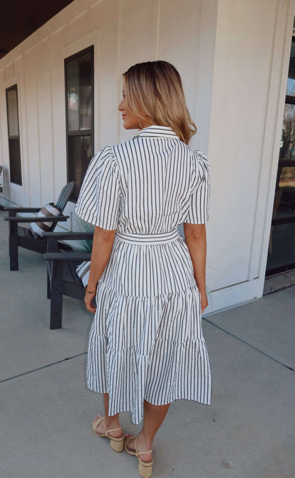 Woman in a White Stripe Surplice Tiered Midi Dress with black stripes stands outside near chairs, facing away from the camera.