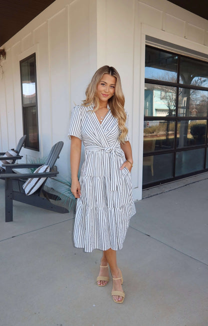 Woman in a White Stripe Surplice Tiered Midi Dress and sandals stands outside near chairs and a modern building.