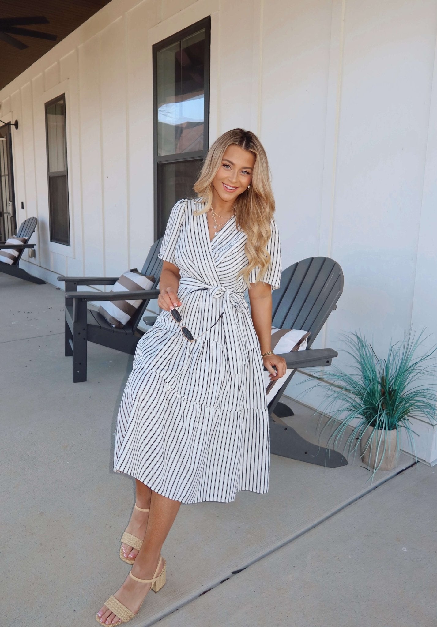 A woman in a White Stripe Surplice Tiered Midi Dress smiles on a porch, holding sunglasses near gray chairs and a potted plant.