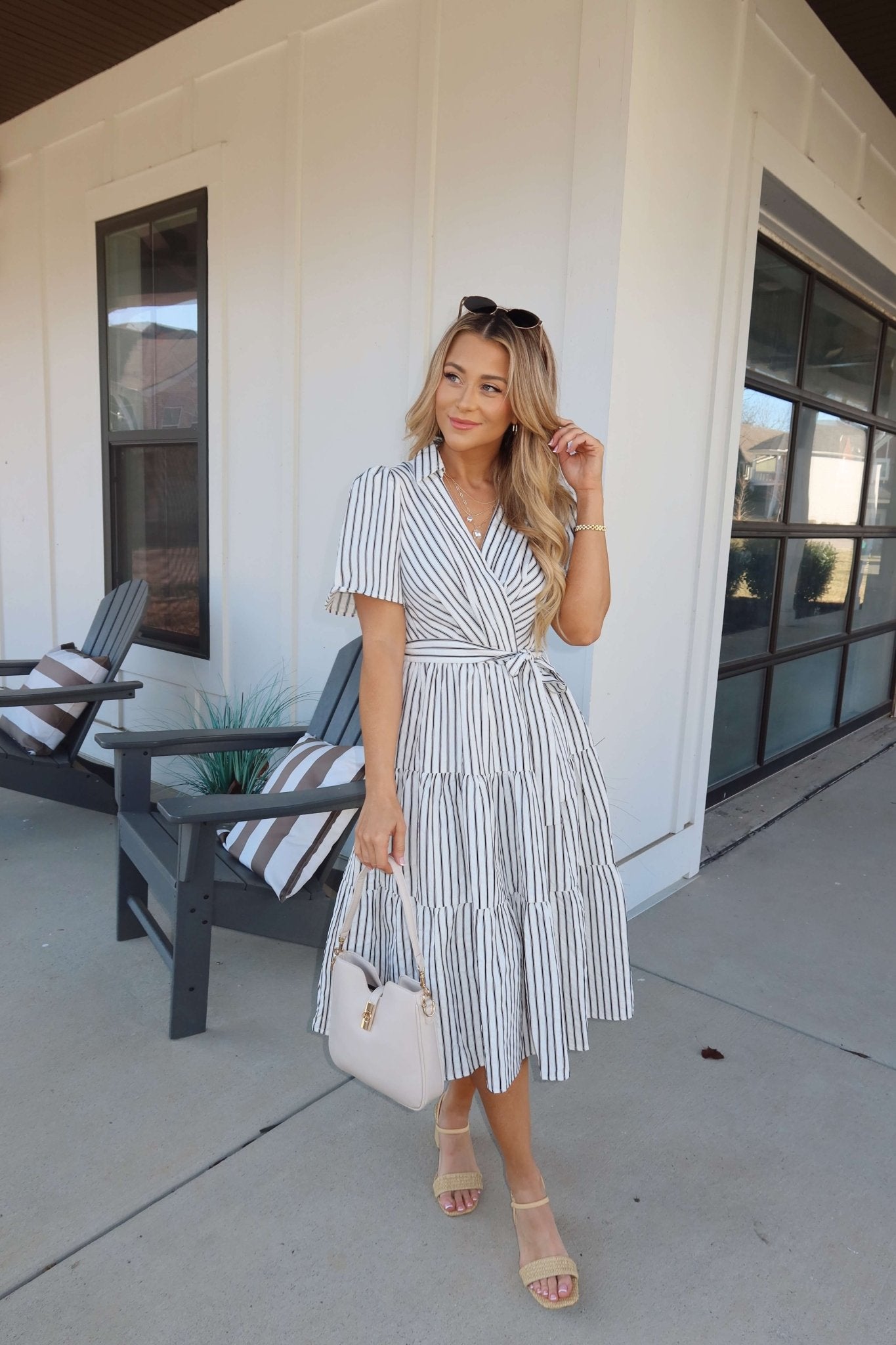 Woman in a White Stripe Surplice Tiered Midi Dress holds a beige handbag on a sunny porch with chairs and a modern building behind her.