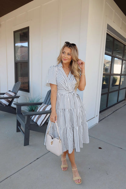 Woman in a White Stripe Surplice Tiered Midi Dress holds a beige handbag on a sunny porch with chairs and a modern building behind her.