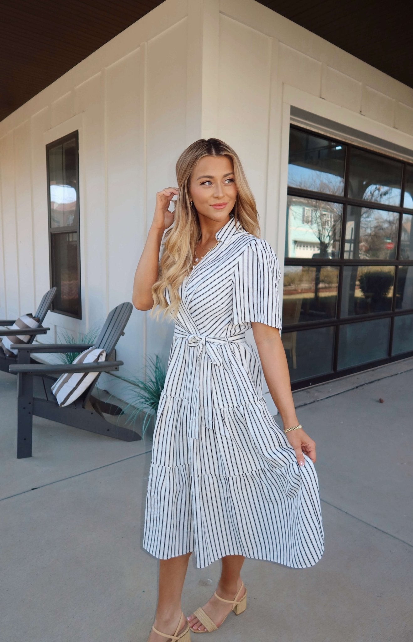 Woman in a White Stripe Surplice Tiered Midi Dress and sandals smiles outside a modern building, touching her hair.