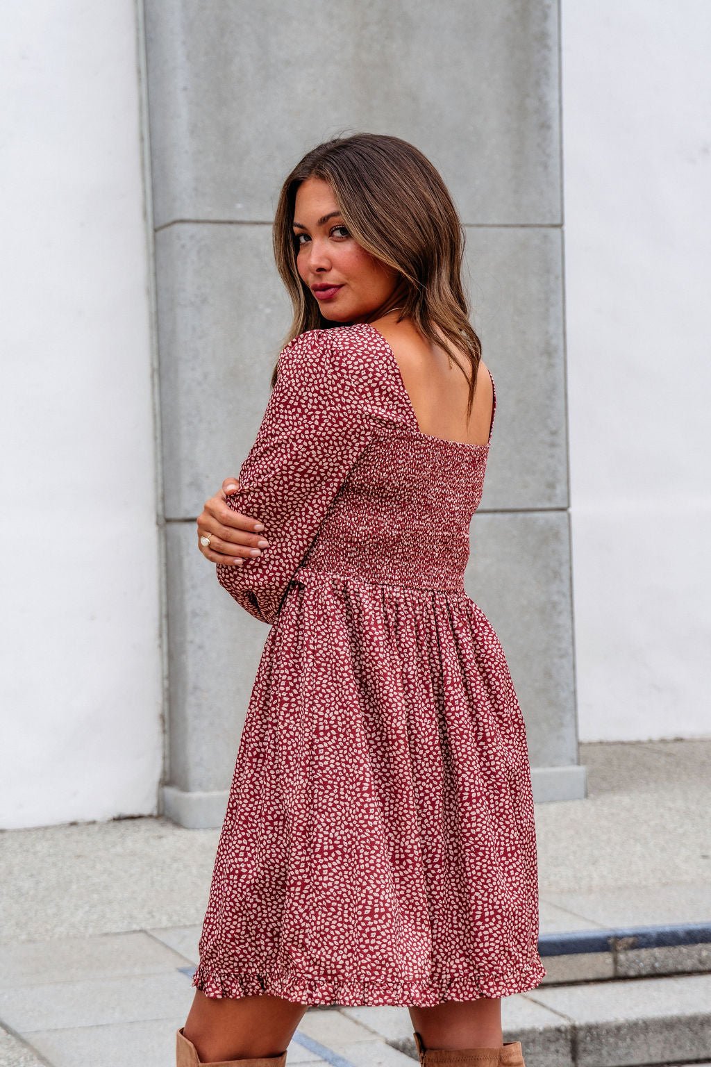 A woman stands outdoors in the Wild Rust Print Smocked Mini Dress - FINAL SALE, looking over her shoulder at the camera.